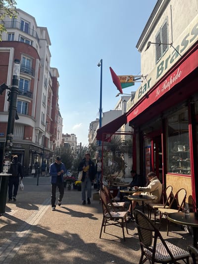 The image depicts a sunny daytime street scene in Asnières-sur-Seine, France. On the right, a commercial establishment identified as "Bar Brasserie Le Rapide" features a red retractable awning extending over an outdoor seating area. Above the awning, a prominent red conical sign with "Tabac" and a bird silhouette is visible, alongside a smaller green sign. The facade is light-colored with large windows showcasing merchandise. Several dark, round cafe tables with woven-back chairs are arranged on the sidewalk; at least two individuals are seated, one in a light-colored jacket appears to be looking down at an object on the table.
On the left side, a multi-story building with a distinctive red brick and white facade featuring balconies and numerous windows dominates the background. A dark lamppost with two luminaires stands prominently in the far left foreground. A blue street pole equipped with a security camera (CCTV) is positioned slightly to the right of the center, between the two main structures. The sidewalk is paved with light and dark blocks, marked by strong shadows cast from overhead elements and pedestrians.
Several people are walking on the sidewalk towards the viewer. A man on the far left wears a dark jacket and carries a white bag. Another man in a blue jacket and dark pants is looking down at his left wrist. To his right, a man in a dark jacket, light pants, and a yellow fanny pack carries a white tote bag. Further back, a woman walks on the sidewalk. Planters with small trees and potted flowers are arranged on the sidewalk between the pedestrians and the cafe tables. The sky is clear blue with minimal clouds. The street extends into the distance, revealing additional buildings.
