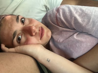 A person lies sideways on white bedding, gazing upwards with a calm expression. Their left arm rests on their forehead, revealing a small tattoo of the letter 'M' on the wrist. They have brown eyes, a nose ring, and flushed cheeks. A light purple sweatshirt is visible on their left side. The background shows blurred skin and clothing, possibly indicating someone lying beside them.