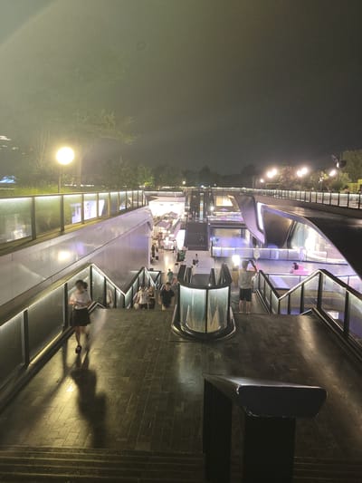 The image depicts an outdoor, multi-level urban scene at night in Wuhou District, China, with a focus on modern architecture and pedestrian activity. The foreground shows a set of dark, textured stairs leading down. To the left, a woman with dark hair, wearing a white t-shirt and a dark skirt, is walking down the stairs, casting a long shadow. Her posture suggests she is moving at a moderate pace.
The midground reveals more stairs and walkways, with metal railings illuminated by integrated lighting. Several people are visible on these walkways, some walking, others standing. One man in the mid-right foreground is standing on a lower level, facing away from the camera, holding a mobile phone and appearing to take a photograph or video. He is wearing a light-colored t-shirt and dark shorts.
Further into the background, the scene opens up to a wider plaza-like area with more contemporary architectural elements, including curved structures and illuminated glass panels. Shops or retail spaces are indicated by the lighting and displays visible through the glass. The overall environment is brightly lit by numerous artificial lights, including street lamps and architectural lighting, which create a vibrant, albeit somewhat hazy, atmosphere. The sky is dark, consistent with nighttime. The architecture is sleek and modern, featuring clean lines and reflective surfaces. The presence of people indicates that the location is a public space, likely a commercial or entertainment district.
