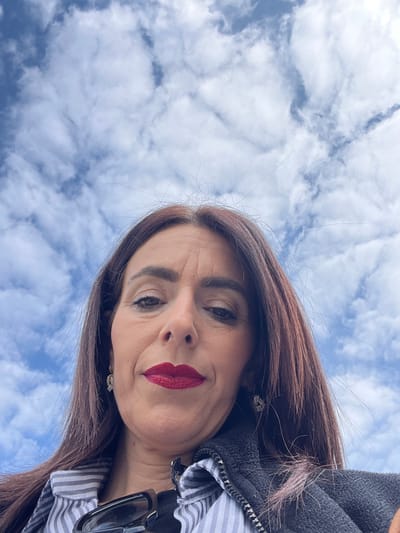 The image displays a female subject, appearing middle-aged, centered against a background of a blue sky scattered with white cumulus clouds. Her dark brown hair, parted in the middle, falls around her shoulders. Visible facial features include wrinkles on her forehead and around her eyes. She is wearing dark red lipstick and has subtle eye makeup, including eyeshadow and eyeliner. Both ears display light-colored, possibly diamond-studded, earrings. She is dressed in a light-colored striped shirt, worn underneath a dark, zippered vest or jacket. Dark-framed sunglasses are tucked into her clothing below her chin. The subject is looking directly at the camera, with the image captured from a low, upward-looking angle. This outdoor scene is located in Roodt-sur-Syre, Luxembourg.