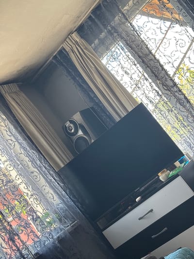 The image is taken from a low angle looking up at a window covered with lace curtains. Behind the lace curtains are heavier, solid-colored curtains. A window is visible through the lace. To the right of the window, a black and white cabinet is partially visible. Underneath the cabinet and above the curtains, a pair of speakers are mounted on the ceiling.
There is no information available to determine if the image was taken in Novi Pazar, Bulgaria.