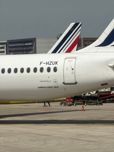 The image shows the rear section of an Air France Airbus A220-300, registration number F-HZUK, parked on the tarmac at a location implied to be in or near Le Mesnil-Amelot, France, given the Air France building and livery on the aircraft. A building displaying the Air France logo with the phrase "S'ENVOLER EN TOUTE ÉLÉGANCE. AIRFRANCE" is visible in the background. The aircraft has an emergency exit door, a tail with blue and white stripes, and another tail with red, white, and blue stripes. Servicing equipment, including what appears to be a refueling or maintenance vehicle, is alongside the aircraft. A person's legs are visible near the servicing equipment.