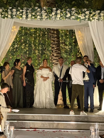 The image captures a wedding celebration, likely outdoors in the Beer Tuvia Regional Council, Israel. The scene is decorated with lush green vertical gardens adorned with strings of warm, fairy lights, creating a festive and intimate atmosphere. A white floral archway, possibly part of a chuppah or a decorative entrance, frames the gathering.
A group of people are the main subjects. Towards the top of the frame, several men are dressed in suits and shirts, some appearing to be in conversation or posing for photos. A man in a white shirt and dark pants holds a camera, suggesting a photographer or a guest capturing the moments. Below them, a woman in a white wedding gown stands out, her expression joyful. Next to her, another woman wears a black, shimmering dress, and further down, other women are dressed in elegant evening wear. The presence of a wedding dress and the attire of the guests strongly indicate a wedding ceremony or reception is underway.
The lighting suggests it is evening or nighttime, with the fairy lights providing a soft glow. The overall mood seems celebratory and warm, with smiles visible on some faces, particularly the bride. The combination of natural elements like the greenery and the soft artificial lighting creates a romantic ambiance. There is no visible text in the image.