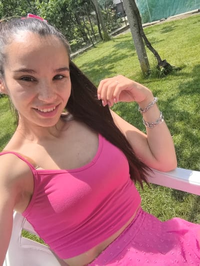 A young female subject is depicted from the mid-chest up, seated outdoors in a white plastic chair in Kran, Bulgaria. She has long, dark brown hair pulled back into a high ponytail, secured with a visible pink hair tie. Her skin is fair with numerous freckles across her nose and cheeks. She has brown eyes and a wide smile, revealing her upper teeth. She wears a bright pink sleeveless crop top with thin straps and a matching bright pink skirt with a textured, dotted pattern. Her left hand is partially visible, featuring a silver ring on her ring finger and pink nail polish. Her right arm rests on the chair's armrest, adorned with two silver-toned chain-link bracelets on her wrist. The background features a vibrant green lawn under bright sunlight, casting shadows on her clothing and the grass. Behind her, various trees and foliage are visible, along with a light green mesh fence or netting (possibly a sports court barrier) in the mid-ground right, and a dark, thinner metal fence further to the left. A prominent tree trunk with a smaller, gnarled branch extending from its base is visible to the right of the subject.