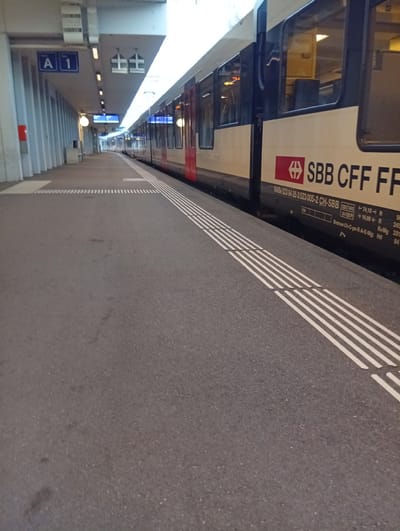 The image depicts a train platform in Langenthal, Switzerland. A train is stopped at the platform, with its doors closed. The train has "SBB CFF FFS" branding on its side. The platform is paved with asphalt and has tactile paving strips for visually impaired individuals. Overhead, there are signs indicating "A 1" and other directional information. The environment appears to be a modern train station, with a covered walkway extending along the platform. The lighting suggests it could be daytime or evening.