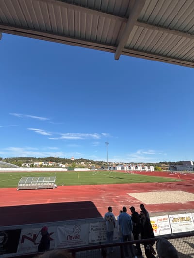 The image depicts a track and field event viewed from stadium seating. On the right side of the frame, several people are visible, with some facing away and others obscured. Signage is attached to the wall with the name “Juvenal Padaria E Pastelaria, Polar Tradicional.” Other signage displays contact information and business details. The track area consists of red running lanes and a green field, and an awning-like structure is at the corner of the track. In the background, buildings and a townscape are visible. Above the townscape is a clear blue sky with sparse clouds. The image seems to be taken in Valpaços, Portugal, based on the business signs visible. A metal stadium overhang is on the left side of the frame.