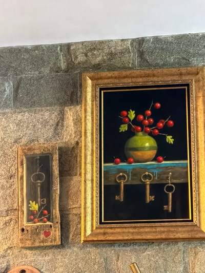 The image shows two framed artworks hanging on a stone wall. The larger artwork features a green and brown striped object with red berries and three keys against a dark blue background. The smaller artwork depicts a key, berries, a green leaf, and a red heart. The image might have been taken in Кирково, Bulgaria, but there is no confirming evidence within the photograph.