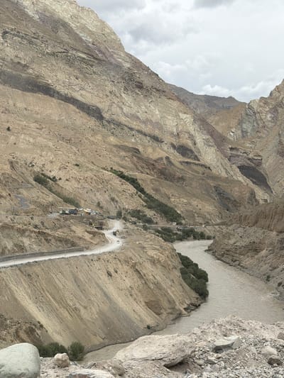 A wide-angle, high-angle shot captures a panoramic view of a mountainous landscape near Yulchung, India. A muddy, fast-flowing river cuts through a deep canyon. On the left side of the canyon, a winding road is carved into the steep, tan-colored mountainside. Several buildings, possibly a small village or settlement, are visible along the road. Trucks and cars are present on the road. The mountains are rugged and appear to be composed of sedimentary rock layers, with varying shades of tan, brown, and some greyish-purple hues. The sky is overcast with grey clouds. The overall impression is one of a remote and arid mountainous region.