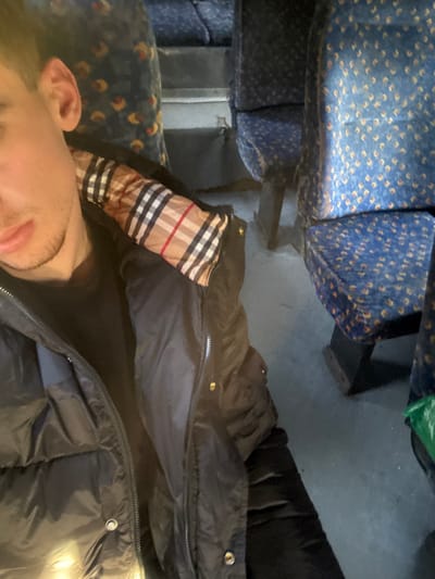 A cropped, slightly high-angle photograph captures a male individual seated within a public transport vehicle. The subject occupies the left side of the frame, showing his profile from the lower lip to the upper chest. He has short brown hair, a visible right ear, and light stubble on his jawline. He is wearing a dark, possibly black, puffer jacket with the main zipper partially open, exposing a black inner garment and a beige, black, and red plaid lining at the collar. To the right and background, multiple rows of blue upholstered seats are visible, each featuring a repeating pattern of small yellowish-orange dots. These seats are supported by dark, solid bases. The floor is a uniform light grey. A small fragment of a green object is discernible on the floor in the bottom right corner, adjacent to a seat base. The setting is consistent with the interior of a bus or train. No specific visual identifiers confirming the city of Partskhanakanevi, Georgia, are present in the image.