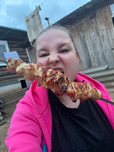 A close-up, outdoor, vertical, medium shot shows a young woman with a silver ring in her nose, wearing a bright pink hooded jacket over a black shirt with small silver dots, about to bite into a skewer of cooked chicken. The chicken is golden brown and appears grilled. Her eyes are squeezed shut and her face is contorted in a humorous expression. The background shows a rustic wooden building with a corrugated metal roof and some laundry hanging on a clothesline. The lighting suggests it is late afternoon or early evening, with overcast skies. The location is identified as Baldeyka, Russia.