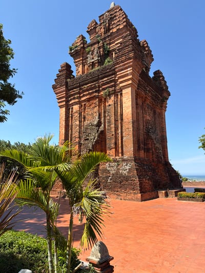 A tall, weathered, red brick tower stands prominently under a clear blue sky in the city of Tuy Hoa Ward, Vietnam. The tower, which appears to be ancient, exhibits signs of age with moss and vegetation growing on its brickwork. It has a tiered, pyramidal structure with intricate architectural details and is surrounded by a large, red-tiled courtyard. In the foreground, lush green palm fronds fan out, partially obscuring the view of the tower and courtyard. A person is visible in the distance, standing near the tower. The environment suggests a warm, sunny day with a glimpse of the ocean in the far background.