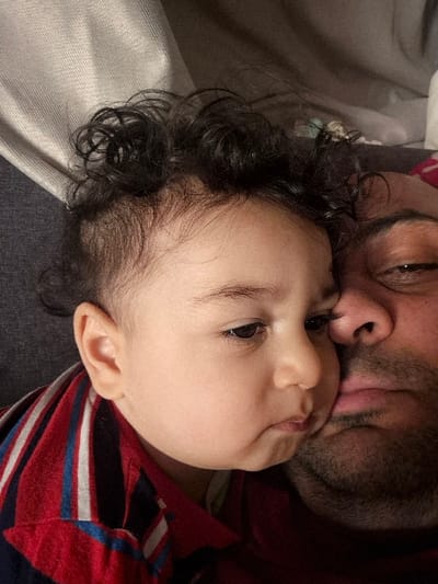 An adult male and an infant are depicted in a close-up, indoor scene located in Melle, Belgium. The infant, positioned on the left side of the frame, has dark curly hair, fair skin, and is wearing a red shirt with blue and white stripes. Its face is pressed against the right side of the adult male's face, specifically contacting his nose and upper lip area. The adult male, visible on the right, has dark facial hair and is wearing a dark-colored shirt; his right eye is partially open and appears heavy-lidded. The background consists of soft, indistinct textile surfaces, suggesting an interior setting.
