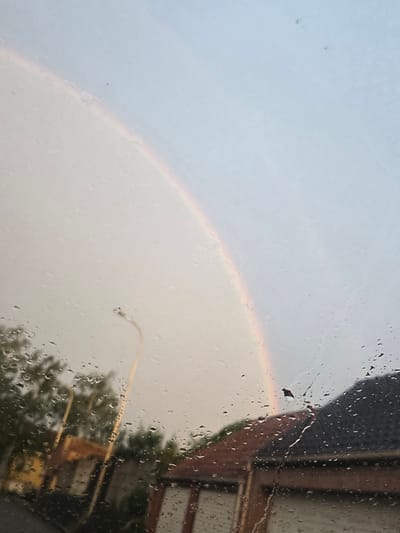 The image presents an outdoor scene viewed through a rain-streaked window, from an apparent indoor vantage point. Numerous water droplets of varying sizes cover the glass, creating a blurred and distorted perspective of the exterior.

The most striking feature is a partial rainbow, visible as a faint arc of color spanning from the middle to the lower-left portion of the frame. The sky beyond the window appears overcast and light, with hints of grey and pale blue, suggesting recent or ongoing rainfall coupled with sunlight breaking through.

Through the rain-covered glass, details of the surroundings are discernible. In the upper right quadrant, a section of a building is visible, characterized by a dark, possibly tiled or shingled roofline or overhang, and a lighter, textured facade that might be brick or wood. A small piece of debris, possibly a leaf, clings to the window in this area.

In the lower right, a distinct, curved, yellow-orange object, consistent with the arm of a street lamp, is apparent. Beneath and around this, blurred dark green shapes indicate the presence of trees or dense foliage.

The scene conveys the atmosphere of a rainy day, likely during the late afternoon or early evening given the formation of the rainbow. No people are visible, and there is no discernible text in the image. The specified location for this image is Mouscron, Belgium.