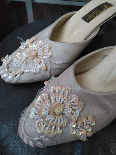 The image displays a pair of light-colored, embellished women's slip-on shoes, or mules, resting on a dark, indistinct surface. The shoes are crafted from a textile material, possibly satin or silk, in an off-white or champagne hue. Both shoes feature intricate embroidery, beadwork, and sequins arranged in floral patterns on their toe boxes. The sequins are small, iridescent, and appear in shades of pink, peach, and gold, while the beads are small, spherical, and off-white. Some loose threads are visible among the embellishments, particularly on the left shoe. The interior sole of the right shoe shows a black label with "Melati" in white text. The shoes exhibit minor signs of wear, particularly on their footbeds. No people are visible. The environment consists only of the shoes on the dark surface; no other objects, actions, or interactions are observable. The image itself provides no visual information to determine a specific location within the city of Kruibeke, Belgium.