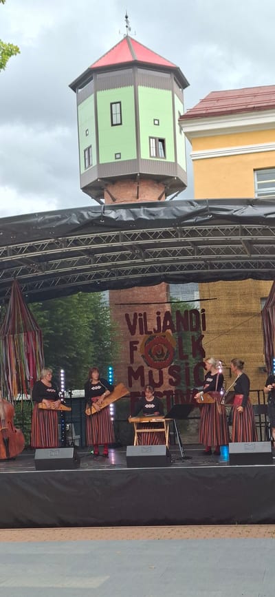 A group of women are performing on an outdoor stage in Viljandi, Estonia. The women are dressed in traditional striped dresses and are playing stringed instruments. Behind them is a large banner that reads "Viljandi Folk Music Festival". In the background, a distinctive green and red water tower is visible, which is a landmark in Viljandi. The stage is set up with lighting and sound equipment. The ground in front of the stage is paved. The sky is overcast.