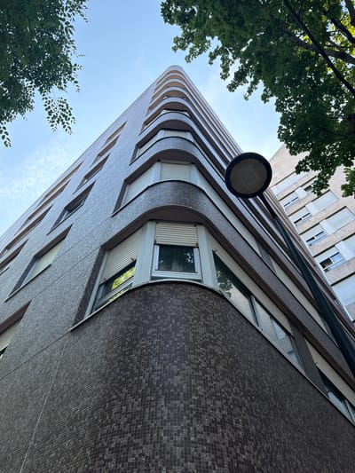 An upward-angle shot captures a building with a curved facade covered in dark brown mosaic tiles, accented by horizontal white bands and numerous windows with closed roller shutters. A tall black lamppost with a circular fixture stands in the foreground, partially obscuring the view of the building. In the upper right and left corners, green tree branches with visible leaves extend into the frame, reaching towards a pale blue sky with faint white clouds. The building's architectural style suggests a mid-20th-century design. Another building, with a lighter, textured facade and similarly shuttered windows, is visible further in the background. The scene is set in Courbevoie, France, likely in a residential or commercial area.