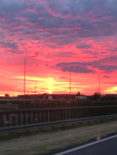 This is a photograph of a vibrant sunset over Srocko Małe, Poland. The sky is dominated by dramatic hues of pink and yellow, with streaks of purple and blue from the clouds. In the foreground, a highway is visible, with a dark barrier separating it from a grassy area. In the middle ground, there are buildings and trees, suggesting a rural or semi-rural setting. Several tall structures, possibly communication towers or light poles, are silhouetted against the bright sky. The overall impression is one of natural beauty and the peaceful end of a day.
