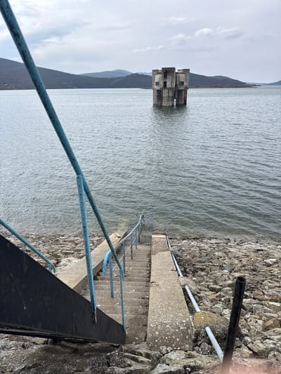 The image depicts a concrete staircase and rocky shoreline leading into a large body of water, identified as a reservoir near Баня, Bulgaria. In the midground, a partially submerged, multi-story concrete structure with exposed openings stands isolated within the water. The foreground features a set of concrete steps descending towards the water's edge, bordered by faded blue metal handrails on the left and a concrete ramp or slab on the right. A dark, slanted metal beam is visible on the far left. The shoreline consists of varying sizes of grey rocks and rubble, with sections of pipes running along the ground near the water. The water surface shows slight ripples. In the background, rolling mountains with sparse, dark vegetation are visible under an overcast, light grey sky. There are no people present in the scene, and no discernible actions or interactions are occurring.