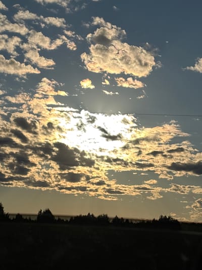 The image depicts a silhouetted landscape under a dramatic sky. The foreground shows a dark, indistinct area that appears to be fields or ground cover, with a line of trees and foliage forming a dark horizon. Above this, the sky is filled with a multitude of clouds. The sun is partially obscured by these clouds, casting bright, golden light that illuminates the edges of the clouds, creating a textured and dynamic effect. The sky transitions from a lighter, warmer hue near the horizon to a deeper blue higher up. Two thin, dark horizontal lines, possibly power lines, are visible across the upper portion of the sky. The overall scene evokes a sunset or sunrise. There are no people or discernible actions. The location is given as Yartsevo, Russia.