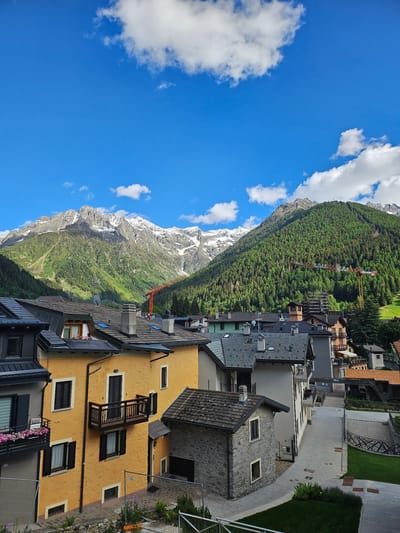 This image presents a picturesque, sunlit view of a mountain town, identified as Ponte di Legno, Italy. The scene is set against a dramatic backdrop of towering mountains, some of which feature snow-capped peaks that contrast with the vibrant green forests covering their lower slopes. The sky above is a clear, brilliant blue, punctuated by scattered white, cumulus clouds.

In the foreground and mid-ground, a collection of buildings forms the heart of the town. These structures showcase a variety of architectural styles, blending traditional elements such as stone facades and wooden balconies with more modern designs. A prominent warm yellow building with dark windows, a wooden balcony, and planters filled with pink flowers stands out in the immediate foreground. Other buildings exhibit dark gray or black sloped roofs, often featuring dormer windows. Amidst the verdant forest on the mountainside, a large red and white construction crane is visible, indicating ongoing development. Paved paths and neatly maintained grassy areas can be seen between the buildings.

The bright and even illumination suggests it is a clear day, likely morning or early afternoon. The weather appears calm and sunny. No people are visible, lending a serene atmosphere to the mountain landscape. There is no visible text in the image.