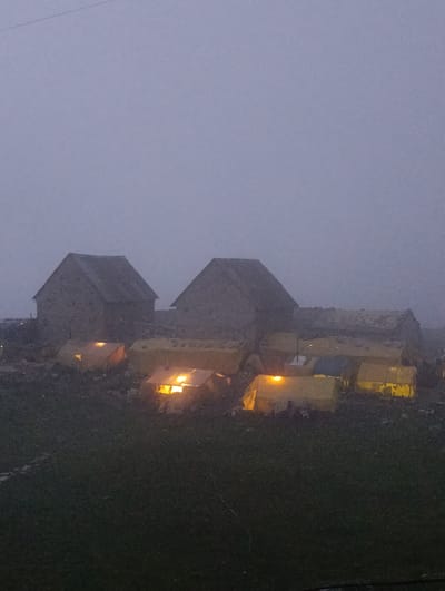 The image depicts a dimly lit, misty scene of a settlement in Marhi, India. In the foreground, a field of dark green grass stretches across the lower portion of the frame, with faint traces of what may be pathways or boundaries. In the middle ground, a cluster of tents are visible, many illuminated from within by warm, yellow light, suggesting occupancy or interior lighting. The tents appear to be of a practical, possibly camping or temporary dwelling design, in various shades of yellow and possibly blue. Behind the tents, two rustic, stone or dark-colored buildings with steep, weathered roofs are silhouetted against the hazy sky. The overall atmosphere is one of subdued light and a pervasive mist, typical of high-altitude environments or certain weather conditions. The image captures a quiet, still moment in what appears to be a remote, possibly mountainous, location.