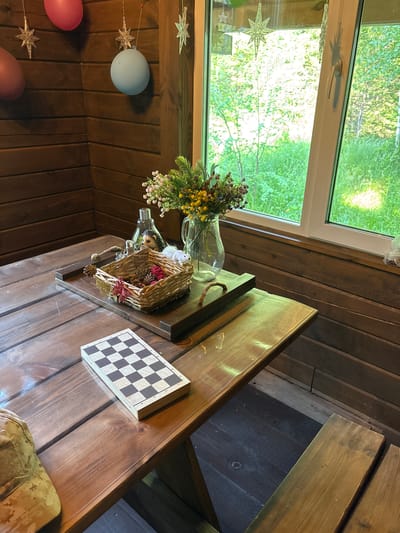 A wooden picnic table sits in a rustic room with wood-paneled walls. On the table, a chessboard is laid out, with a small basket of decorative items and a vase of flowers nearby. Through a large window, lush green trees and foliage are visible, suggesting an outdoor setting. The room appears to be part of a cabin or a similar rural structure, possibly located in the village of Bol'shaya Rechka, Russia, given the context. There are no people present in the image.