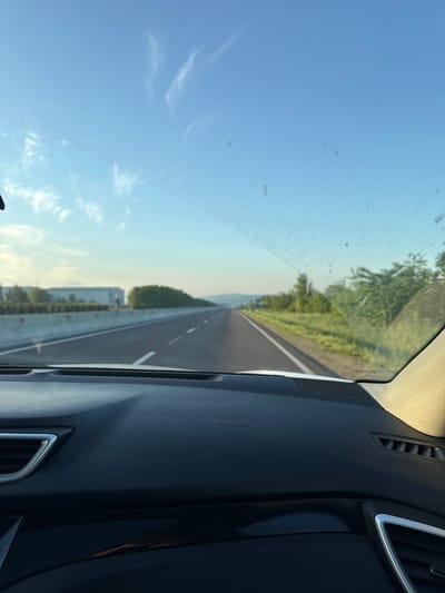The image is a first-person view from inside a car driving on a highway in Cojușna, Moldova. The road stretches into the distance, flanked by green fields and trees on either side. A concrete barrier runs along the left side of the highway, with a large building and more trees visible beyond it. The sky is clear blue with a few wispy clouds. The car's dashboard is visible in the foreground, with the words "SRS AIRBAG" printed on it.