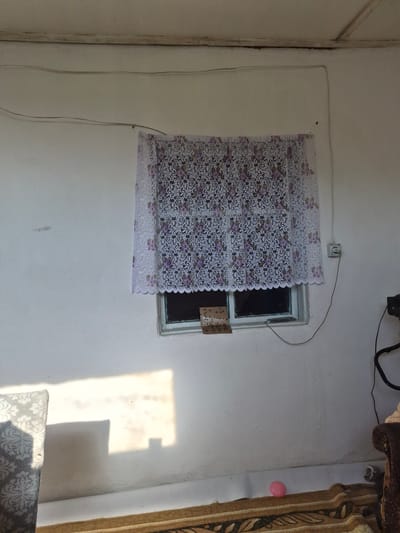 The image depicts an interior scene, possibly a room with a window. The walls are painted white, and there are some electrical wires running along the ceiling and down the wall. A window is covered with a white lace curtain with a floral pattern in purple and green. The window itself has a simple frame. Attached to the outside of the windowpane is a small piece of paper, possibly a note or advertisement. Sunlight is casting a bright rectangle of light onto the wall to the left of the window, and also onto a portion of a patterned sofa visible in the lower left corner. A section of a patterned rug is visible on the floor. In the lower right corner, a portion of a dark, upholstered furniture item is visible, and on the floor near it is a small pink ball. The overall impression is of a simple, possibly rural interior. No specific location in the city of Қанабек, Kazakhstan can be determined from the image alone.