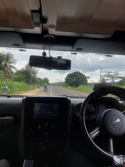 The image displays a driver's perspective from inside a Jeep vehicle traveling on a two-lane asphalt road in Kisanga, Tanzania. The interior foreground features a black dashboard, a central infotainment system showing "BLUETOOTH AUDIO," and a black steering wheel with a "Jeep" logo, partially gripped by a dark-skinned hand. Overhead, a rear-view mirror with an integrated screen displaying a scenic view is visible, accompanied by visible wiring.
The exterior scene shows a paved road extending into the distance, bordered by dense green vegetation, including tall grasses, bushes, and trees; a palm tree is visible on the left. The sky is overcast. On the left road shoulder, an individual wearing a light-colored shirt walks away from the camera. A person operating a light-colored motorcycle is visible in the left lane, also moving away. Further down the road, indistinct figures and vehicles are discernible. Utility poles are present along the right side of the road.