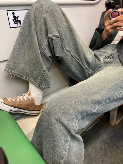 The image shows a person sitting with their legs crossed, wearing light blue wide-leg jeans, white socks, and brown Converse sneakers. The person is holding a cell phone in their left hand. The person is also wearing a gray blazer. A wheelchair accessibility symbol is visible on a white surface to the left. A green seat is also visible. The location is indeterminable without additional information.