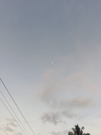 A wide-angle, slightly low-angle shot shows a sky with muted blue and gray tones, suggesting either twilight or overcast conditions. The visible portion of the moon is a small crescent in the upper center of the frame. Wispy clouds are scattered throughout the sky, more concentrated towards the bottom. In the lower right corner, the top of a palm tree is visible. In the lower-left portion of the image, there are three parallel power lines running diagonally from upper right to lower left.

The location is Sumbokrayek, Indonesia, as stated in the prompt. No people are visible, and there is no indication of specific actions or interactions.