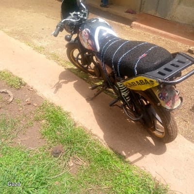 The image displays a black motorcycle, equipped with yellow wheel rims, a black mesh seat cover, and a black rear cargo rack, parked on a light-colored paved surface within Bonyamaro ward, Kenya. A black helmet rests on the handlebars. The motorcycle's fuel tank features white graphics and additional imagery, while the rear fender bears multiple stickers and the yellow text "KABABA". To the left of the paved area, a strip of green grass interspersed with reddish-brown soil contains a small dark rock and a curved, dark object. Beyond the paved surface, a reddish-brown dirt road extends into the background. The lower legs of two individuals, one in blue trousers, are partially visible in the far background near a light-colored structure. The scene is illuminated by daylight, casting shadows from the motorcycle towards the right side of the frame.