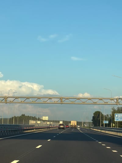 The image depicts a highway scene in Zimnyaya Gorka, Russia, under a clear blue sky with scattered clouds. The perspective is from a moving vehicle, looking forward. In the foreground, a dark asphalt road with white lane markings is visible. A metal guardrail lines the right side of the road.
In the middle ground, several vehicles are traveling on the highway, including cars and a truck. A large overhead gantry structure with metallic supports spans across the highway. Streetlights are positioned along the roadside. In the distance, a dense line of trees forms the horizon.
On the right side of the highway, a blue directional sign with white text and symbols is prominently displayed. The text on the sign appears to read "ОРЕНБУРГ" (Orenburg) and "ТУРГАЙ" (Turgay), indicating directions to these locations. Other smaller signs are also visible in the distance. The overall environment suggests a typical highway setting outside of a populated area, likely during daytime.