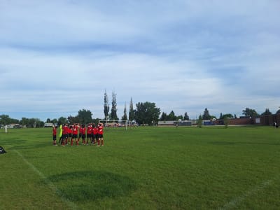 A group of young soccer players in red uniforms are gathered on a grassy field, stretching. The field has white markings for a soccer game. In the background, there are trees, buildings that appear to be part of a school, and a soccer net. The sky is partly cloudy.