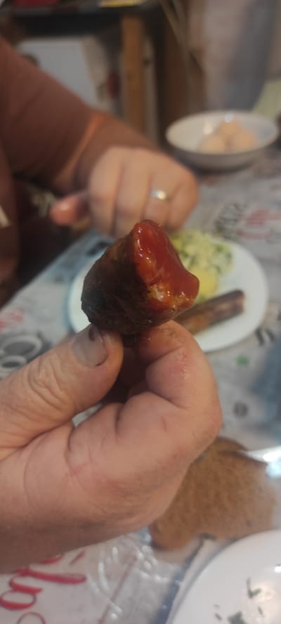 In Kobeliachok, Ukraine, a close-up image shows a person's hand holding a piece of food, possibly a sausage, covered in ketchup. The food item is dark brown and appears to be textured, with a shiny red ketchup glaze. The hand holding the food has visible fingernails and skin texture. In the background, slightly out of focus, is a white plate with what appears to be a side dish of mashed potatoes or similar and another sausage. A white bowl with round, light-colored objects, possibly pastries or dumplings, is also visible. The setting suggests a meal or gathering, with a patterned tablecloth and glimpses of furniture.