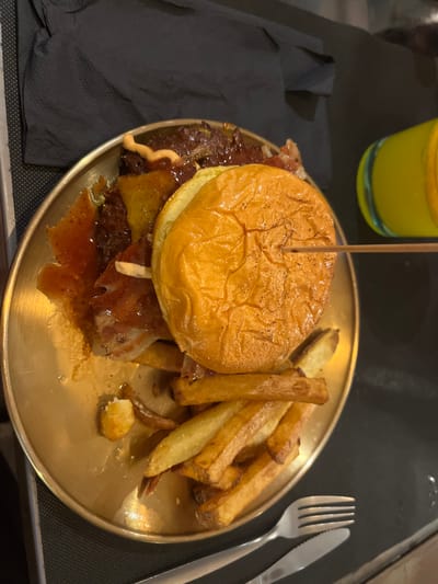 A high-angle shot presents a burger and fries on a golden plate. A wooden skewer pierces the burger's top bun. The burger contains a patty, cheese, bacon, and sauce. Adjacent to the burger are a handful of fries. To the right of the plate, a fork and knife are placed on the surface. The background includes a glass of yellow liquid and a dark napkin. The setting suggests a restaurant environment, possibly in Alcalá de Henares, Spain.
