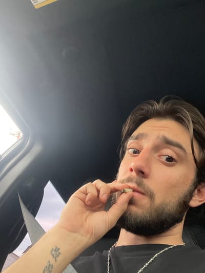 A young man with dark hair and a beard is sitting in the passenger seat of a vehicle, holding a cigarette to his lips. He is wearing a black t-shirt and a silver chain necklace. The interior of the car, including the ceiling and a seatbelt, is visible. The image appears to be taken from a low angle, looking up at the man. Outside the car window, the sky is bright, suggesting it is daytime. There are no identifiable landmarks or clues that indicate the specific location within Ciudad López Mateos, Mexico.