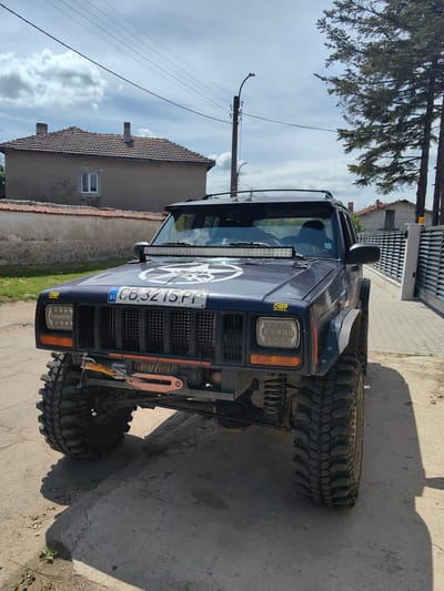 The image depicts a modified Jeep Cherokee XJ parked on a street in the city of Gorni Okol, Bulgaria. The vehicle is navy blue and features numerous aftermarket accessories including large off-road tires, a winch, an LED light bar, and OMP stickers. A Bulgarian license plate is visible reading "CB3215PP". The surroundings include residential buildings, trees, and a street lamp. The setting is a sunny day with some clouds. No people are visible in the image.