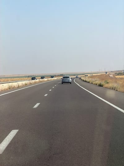 The image shows a gray car traveling on a two-lane road in Nzalat Laadam, Morocco. The road has a white dividing line and white lines marking the shoulder. Other cars are visible in the distance on the left side of the road. The environment consists of dry, open land beside the road. The sky is a pale blue.