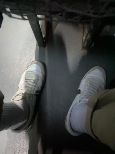 The image shows a low-angle shot of a person's feet wearing white and grey sneakers with white laces. The person is wearing grey socks and beige pants. The shoes are placed on a dark grey, speckled floor, possibly a public transport floor, with a dark metal structure visible in the background. The lighting suggests an indoor or dimly lit outdoor environment. The scene does not contain any recognizable landmarks or features that would place it specifically in Taraclia, Moldova.