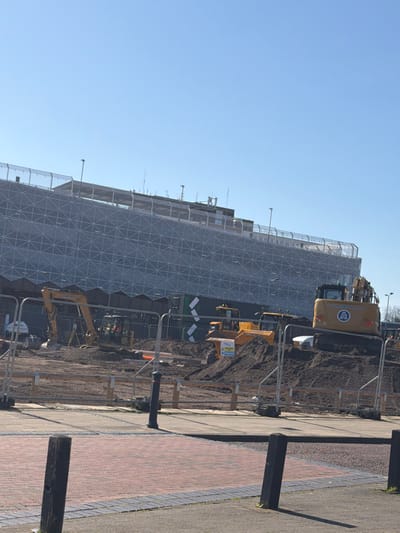 This image depicts a large-scale construction site in Crewe, United Kingdom, specifically located in front of what is identifiable as the Crewe Railway Station Multi-Storey Car Park. The scene is observed from a public pedestrian area under a clear, bright blue sky.
In the background, the multi-story car park rises prominently. Its exterior is covered by a light-colored, patterned mesh facade, featuring a repeating grid of triangular or diamond shapes. The rooftop perimeter is secured by metal fencing, and various small structures, antennas, and light fixtures are visible along its top edge.
The midground is occupied by an active construction zone, enclosed by temporary metal barrier fencing (Heras type) with white plastic feet, and some low wooden barriers behind them. The ground within the site is uneven, consisting of dark brown disturbed earth and multiple soil mounds. Several pieces of heavy machinery are present:
* On the left, a yellow tracked excavator with "SANDERSON" visible on its boom is positioned on relatively level ground. A white tripod-mounted surveying instrument stands nearby.
* Towards the middle-right, a yellow articulated dumper truck is situated atop a mound of earth. A partially obscured white sign, likely a warning, is attached to its front.
* On the far right, a larger yellow tracked excavator, bearing the name "SANDERBROOK" and an 'A' logo within a circle on its rear counterweight, is positioned on a higher mound of soil.
* A large, dark green structure with white diagonal stripes, possibly a container, is partially visible behind the dumper truck. Orange conduits or pipes are visible on the ground.
The foreground shows a paved pedestrian area. In the lower left, the surface consists of reddish-brown brick pavers. Moving right, this transitions to a grey paved surface. Several dark, cylindrical bollards are spaced along the edge of the pavement; one prominent black bollard with a decorative top is centrally positioned in the lower mid-left. No individuals are present in the frame, and no active work is visibly occurring.