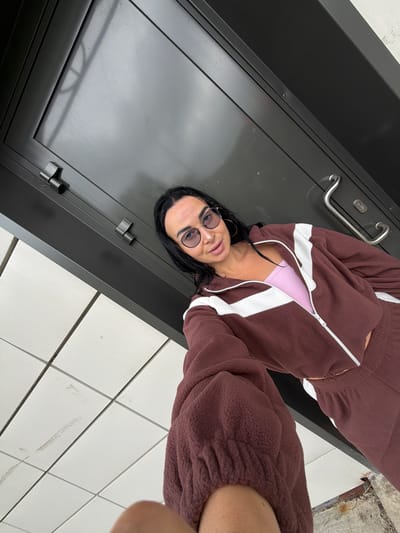 A person with dark hair and glasses is wearing a brown and white tracksuit with a pink top underneath. They are standing on a white tiled floor next to a dark gray or black door. The image appears to be taken at an unusual angle. There is no information in the image indicating the location is in the city of Sust'e-Konets, Russia.
