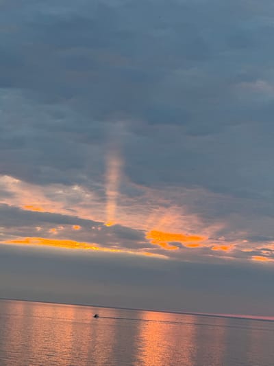 The image depicts a wide expanse of the Baltic Sea at dusk, with the sky dominated by dramatic, dark gray clouds. A shaft of light breaks through the clouds, illuminating a tall, narrow section of the sky with an orange glow. Below, the sea reflects the warm hues of the sunset, creating a shimmering, rose-gold surface. A small boat, the only discernible man-made object, travels across the water, leaving a subtle wake. The horizon line is clearly defined, separating the sea from the sky. The overall impression is one of serene, natural beauty during twilight.