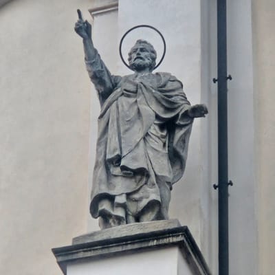 This image shows a stone statue of a bearded man with a halo, standing on a pedestal. The statue is positioned in front of a light-colored, textured wall. The man is depicted wearing draped clothing and has his right arm raised, with his index finger pointing upwards. A dark, vertical pole with two visible fasteners is present to the right of the statue. The statue is likely located in Calcinate, Italy, as indicated by its architectural style and religious iconography commonly found in Italian cities.