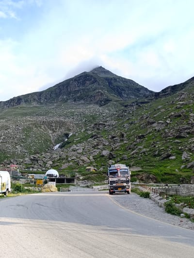 In Damphug, India, on a paved road, a white truck with red and blue detailing is parked. The truck has a license plate that reads "HP 26 C 7777". To the left of the road, a white van is parked. The surrounding environment is mountainous with green grass and large rocks. A waterfall cascades down the side of a mountain in the distance. The sky is cloudy and light blue.