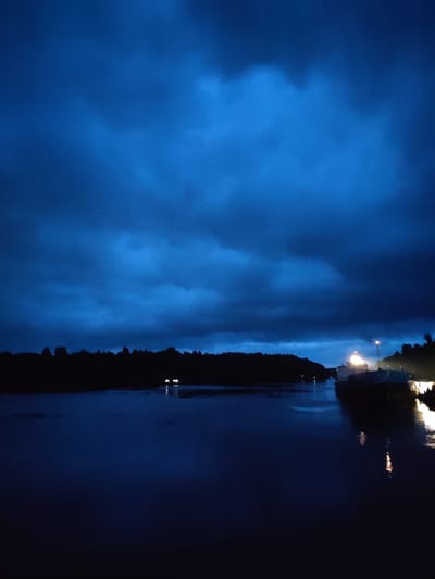 The image depicts a dark, overcast sky with deep blue hues and scattered clouds, suggesting either twilight or a storm approaching. Below the sky, a dark silhouette of a tree line runs across the horizon, separating the sky from a body of water in the foreground. The water is dark and reflects the dim light from the sky and some artificial lights. On the right side of the water, there are two boats docked. One boat is closer to the viewer and appears larger, with visible lighting. Another boat is further away, also illuminated. The scene is dark and quiet, with a sense of isolation. The location is identified as Devyatiny, Russia.