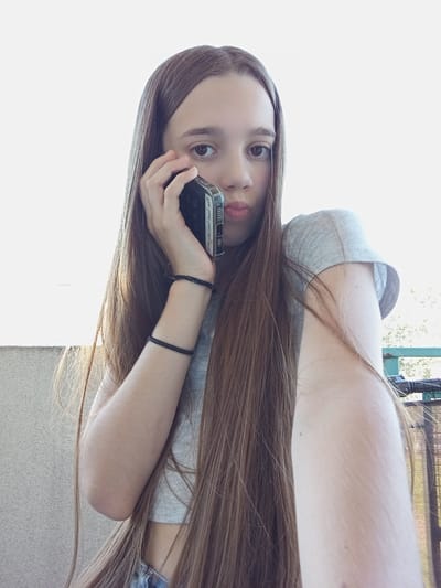 A young girl with long, brown hair is holding a cell phone to her right ear and looking at the camera with her lips pursed. She is wearing a grey crop top. On her left wrist, she has two black bracelets. Her left arm is bent, with her elbow resting on her left shoulder, and her hand is extended towards the camera. She appears to be on a balcony or terrace, with a textured wall to her left and a fence with foliage in the background. The image is taken from a slightly low angle. The bright sky suggests it is daytime. The location is in the city of Radzymin, Poland.