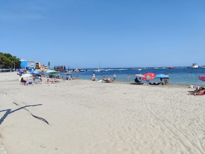 The image depicts a crowded beach in Los Alcázares, Spain. The sandy beach is populated with numerous people sunbathing, sitting in chairs, and wading in the shallow water. Beach umbrellas of various colors provide shade. In the background, the calm blue water of the Mar Menor lagoon is dotted with small boats and a floating structure with a lifeguard tower. Along the shoreline, buildings and trees are visible, suggesting the beach is situated within a developed coastal area. The sky is clear and bright blue, indicating a sunny day.