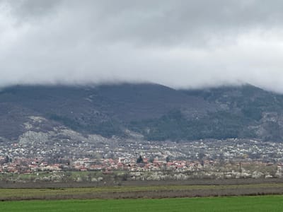 The image depicts a landscape in Manolovo, Bulgaria, on a cloudy day. In the foreground, there is a field of green grass. Beyond the field is a settlement with houses having red roofs. The settlement is interspersed with trees, some of which appear to be in bloom with white flowers. Behind the settlement, a mountain range is visible, partially obscured by low-hanging clouds. The overall atmosphere is overcast.