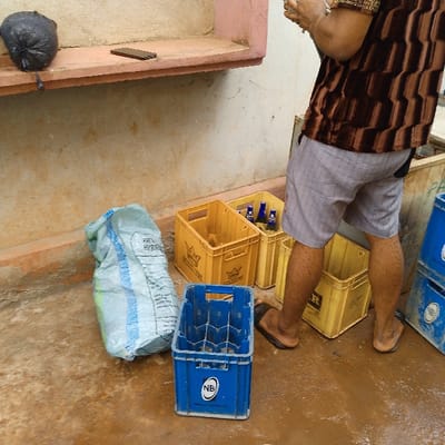 A man stands amongst stacks of beverage crates. The crates, mostly yellow and blue, are scattered around on a wet ground, with a few bottles visible within one. A bag sits near the crates. The man wears patterned brown top and gray shorts, with brown sandals. In the background, a wall has a shelf with a black plastic bag and a phone on it.